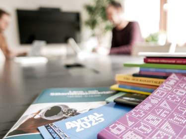 En hög med avtal på ett mötesbord, suddigt i bakgrunden syns en suddig person sitta böjd över en hög papper.