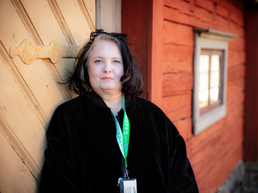 En kvinnlig Seko-medlem på Skansen tittar in i kameran lutad mot en rödmålad trästuga i soldiset.