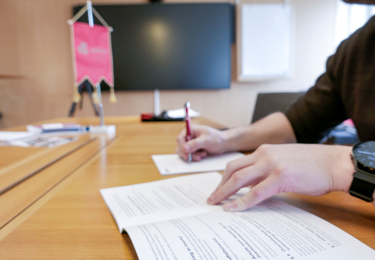 Bild av armar och händer på en person som sitter vid ett mötesbord, gör anteckningar i en avtalsbok. I bakgrunden en Seko-vimpel.