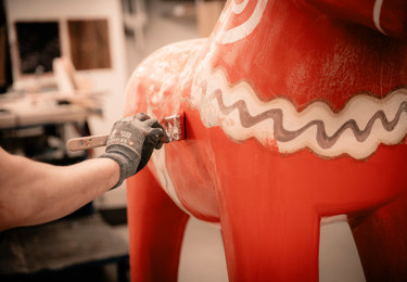 En arm syns i bilden, i handen en pensel som målar på en stor dalahäst.