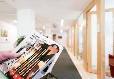 Någon håller en folder med information om Sekos a-kassa i handen, i bakgrunden en korridor med kontor.