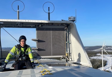 En manlig Seko-medlem i arbetskläder sitter högt upp på en vindkraftverk och tittar in i kameran.