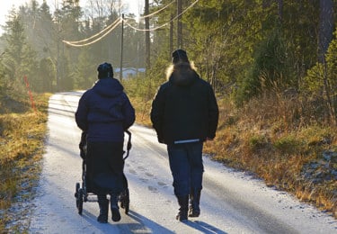Föräldrar med barnvagn på höstpromenad. De är fotade bakifrån på en mindre väg med skog på båda sidor om vägen.