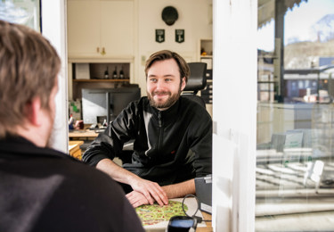 En leende Seko-medlem sitter i biljettkiosken i Skansen entré, i förgrunden en besökare sedd bakifrån.